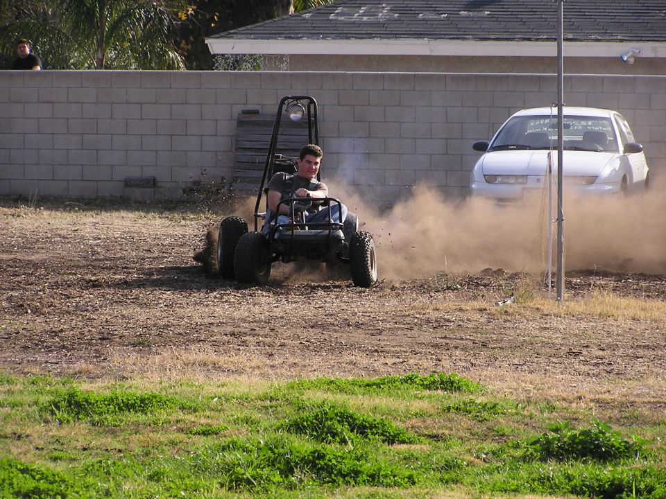 Person driving an off-road go-kart buggy in a dirt yard by a car