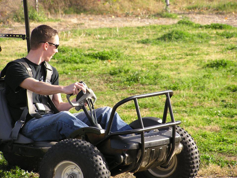 Man in sunglasses driving a tube-frame off-road buggy with knobby tires on grass.