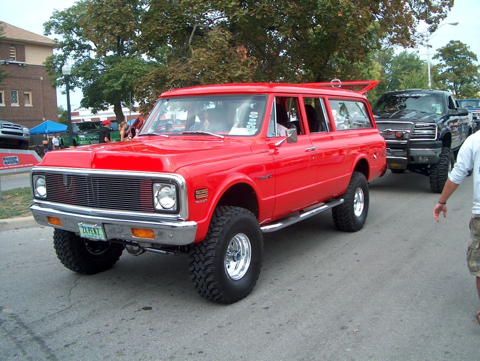 Red lifted Chevrolet Suburban with chrome grille and side steps, followed by a black pickup truck