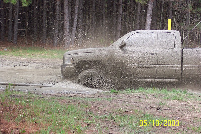 Mud-covered pickup truck splashing through a muddy puddle in a wooded area. Water and mud spray up around the tires and body