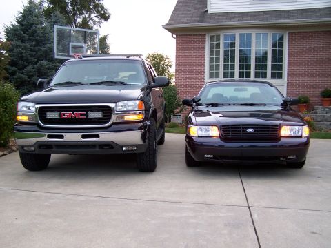 2003 Ford Crown Victoria - Two vehicles, a GMC truck and a Ford Crown Victoria, are parked in front of a brick house.