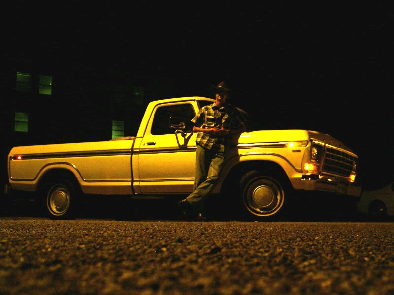 Person leaning on a light-colored older pickup truck at night