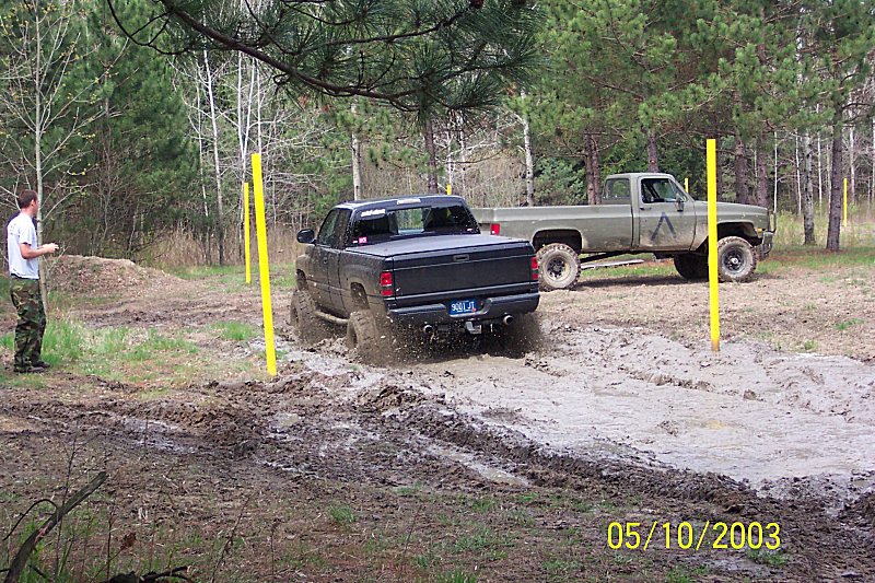 Black pickup truck driving through mud, green pickup truck parked behind it. Person standing to the left