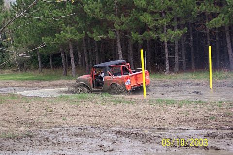 1978 Ford Bronco - An orange off-road vehicle navigates a muddy track surrounded by trees.