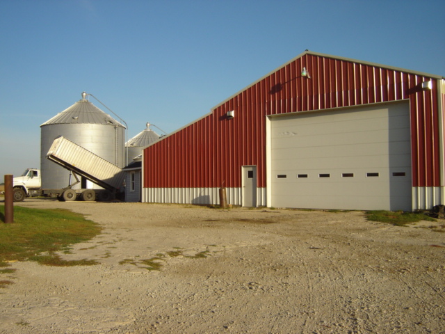 White truck with a trailer backed up to a red barn and grain silos
