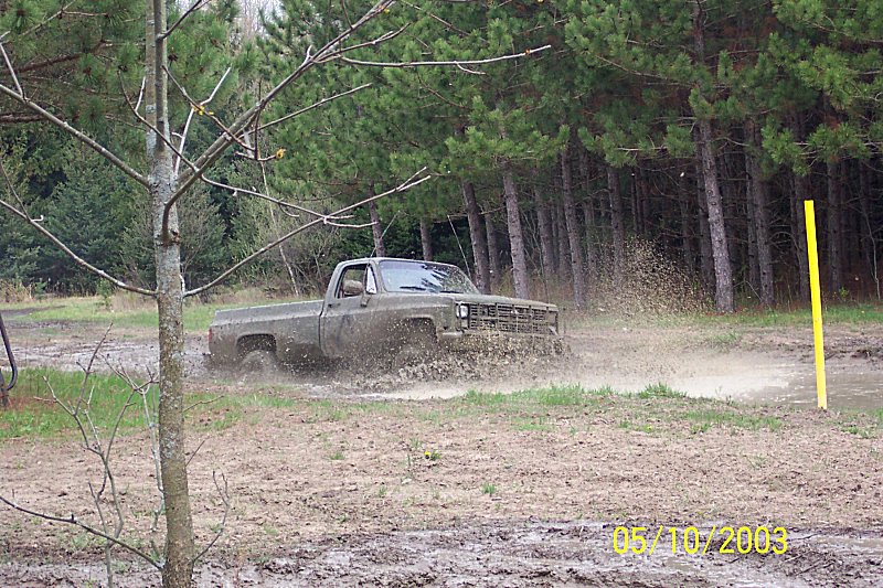 Olive green pickup truck driving through mud, splashing water. Trees and a yellow pole in background