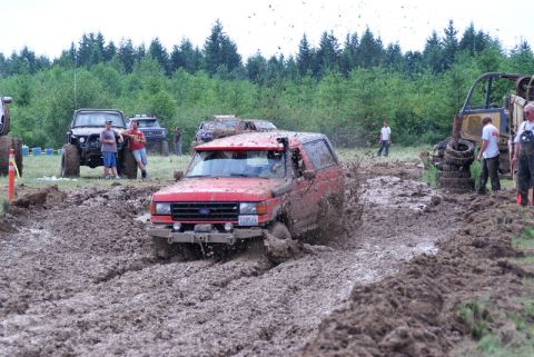 1989 Ford Bronco - A red SUV navigates a muddy track during an off-road event, with spectators and other vehicles present.