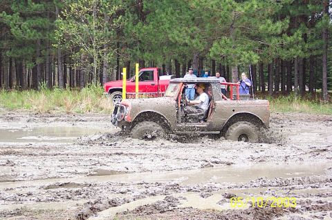 1978 Ford Bronco - An off-road vehicle navigates through a muddy terrain with a red pickup truck in the background.