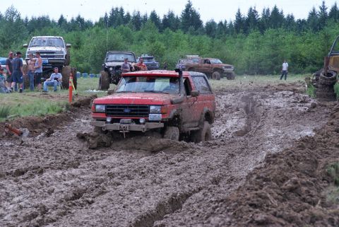 1989 Ford Bronco - A red SUV navigates a muddy track during an off-road event, with other vehicles and spectators in the background.