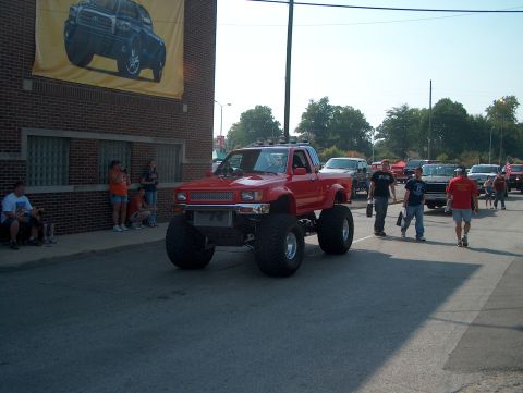 1978 Ford Bronco - A heavily modified red pickup truck with oversized tires is driven down a street, with other vehicles and pedestrians visible.