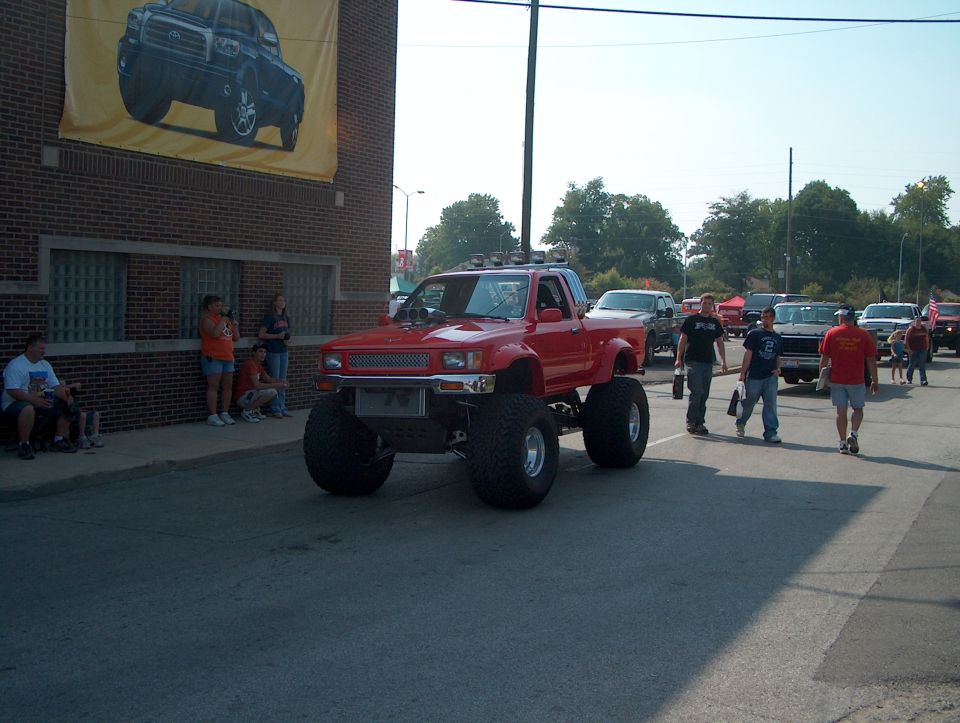 Red lifted pickup truck with large tires, custom grille, and roof lights, seen from a 3/4 front angle