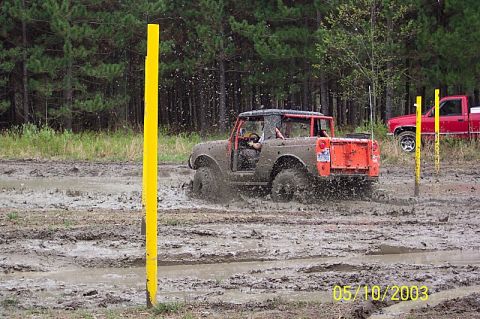 1978 Ford Bronco - A 1978 Ford Bronco navigates a muddy obstacle course, kicking up spray as it goes, with a red pickup truck visible in the background.