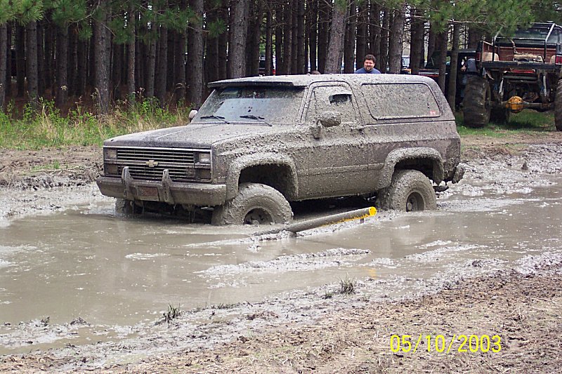 Mud-covered Chevrolet K5 Blazer stuck in mud, front 3/4 view. Visible grille and headlights suggest 1980s model