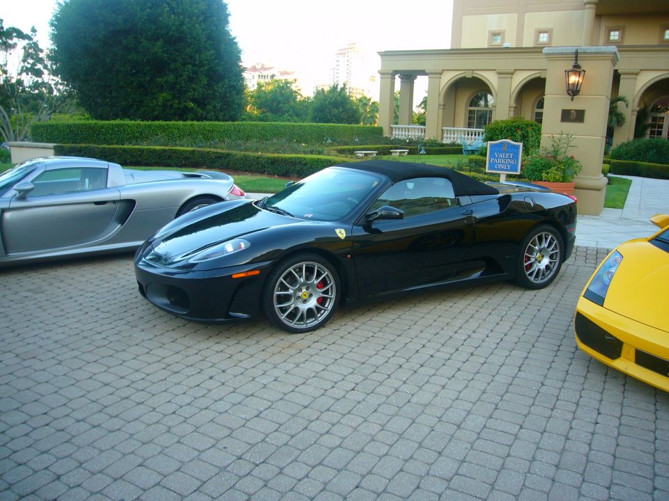 Black convertible Ferrari parked on cobblestones next to a silver Porsche and yellow Lamborghini