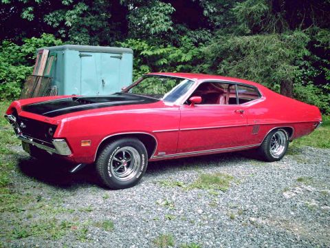 1970 Ford Torino - A red 1970 Ford Torino GT coupe with a black hood sits on a gravel lot, tree-lined lot.