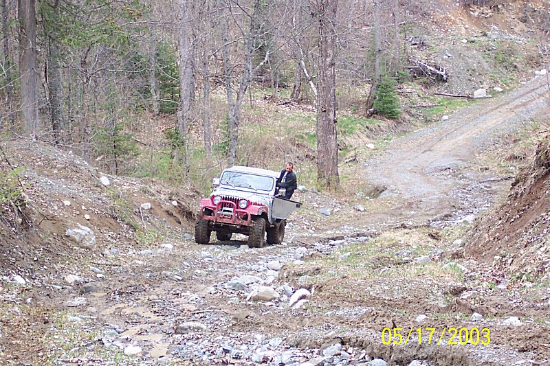 Red Jeep CJ-style 4x4 with windshield down on a rocky forest trail, person standing alongside