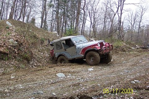 1978 Ford Bronco - A Jeep CJ-7 navigates a muddy, wooded trail, its front tires deep in the muck.