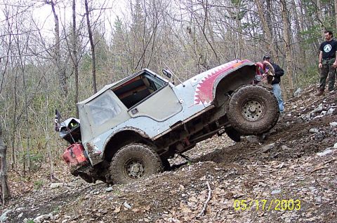 1978 Ford Bronco - A Jeep CJ-7 navigates a steep, wooded trail, its tires struggling for traction on the loose dirt and rocks.