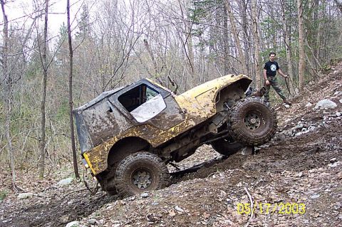 1978 Ford Bronco - A man watches a heavily soiled off-road vehicle navigate a steep, muddy incline in a wooded area.