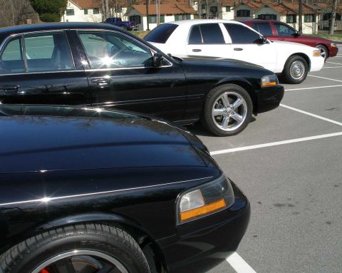 2001 Ford Crown Victoria - Three Ford Crown Victorias are parked in a lot, two black and one white, with a red one in the background.