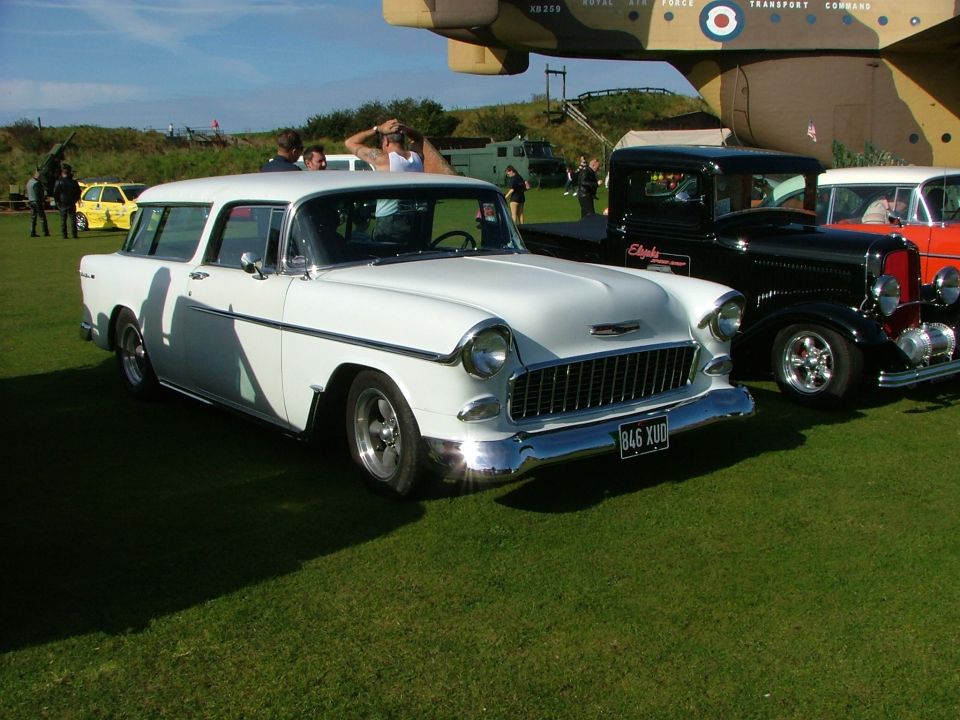 White 1955 Chevrolet Nomad wagon parked on grass with a black pickup and orange car behind it