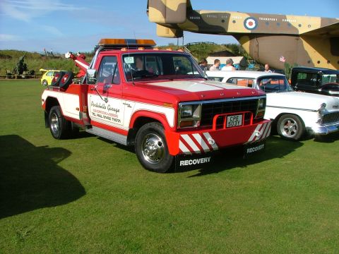 1988 Ford Sport - A red and white tow truck is parked on a grassy field next to other vehicles and a large aircraft.