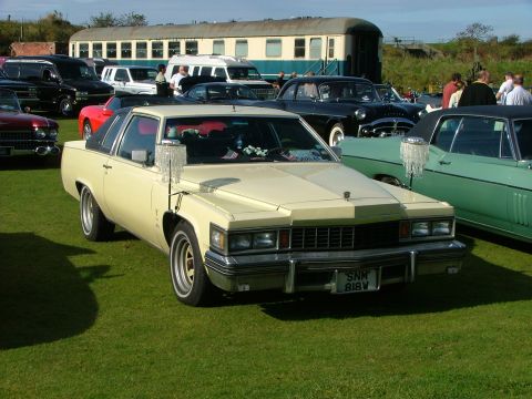 1988 Ford Sport - A cream-colored Cadillac Coupe DeVille from 1977 is parked on a grassy field with other vehicles and a train in the background.