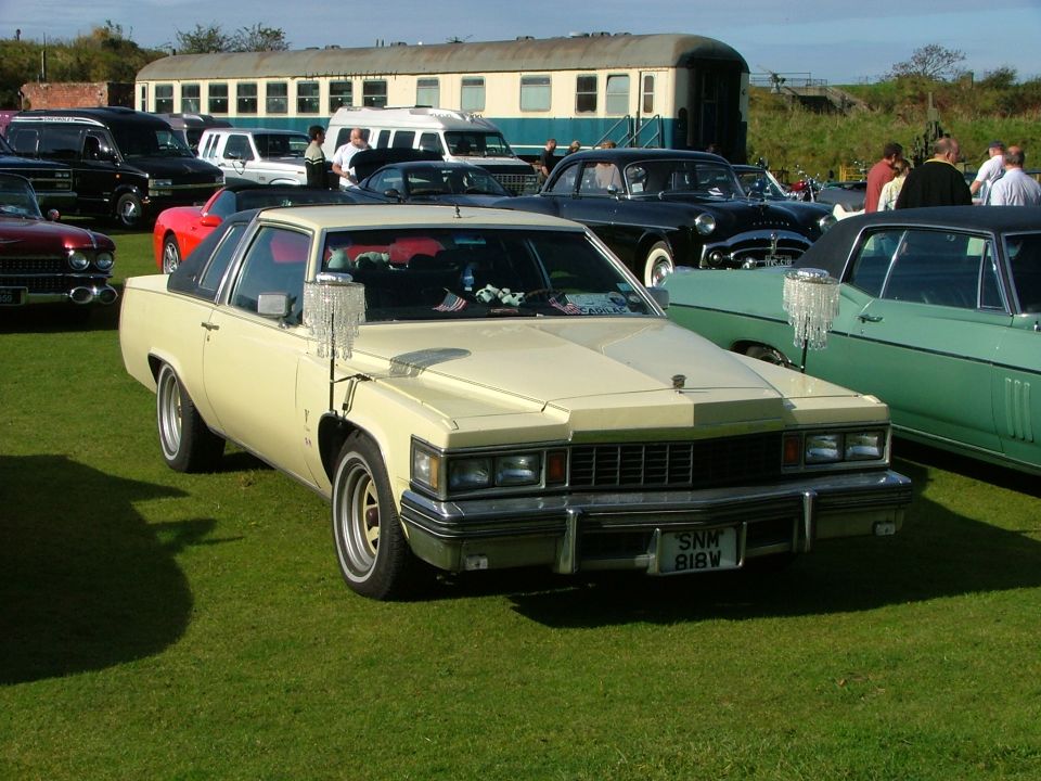Cream 1977 Cadillac Coupe DeVille with a black vinyl roof, parked on grass. Front view shows chrome grille and bumper