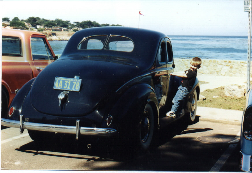 Black 1939 Ford coupe from rear quarter by the ocean, boy climbing on running board, California plate 4A 31 78