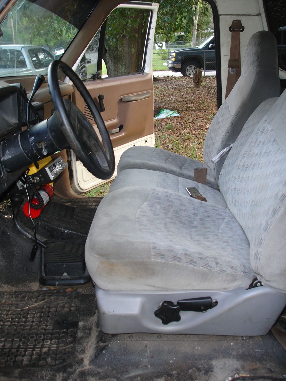 Interior view of a truck showing the driver's seat, steering wheel, and door panel. Another truck is visible outside