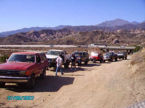1990 Ford Bronco - A line of SUVs and trucks are parked on a dirt road with mountains in the background.