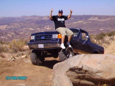 1990 Ford Bronco - A person sits atop a lifted Ford F-Series truck on a rocky, off-road trail with a desert landscape in the background.