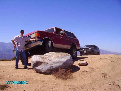 1990 Ford Bronco - A red Ford Bronco is parked on a rock with a man standing beside it, while a dark SUV is visible in the background on a dirt road.