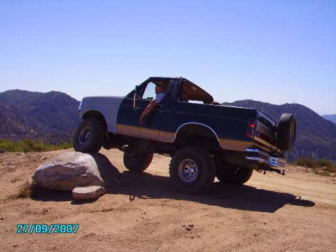 1990 Ford Bronco - A Ford Bronco navigates a rocky trail in a mountainous, arid landscape under a clear blue sky.
