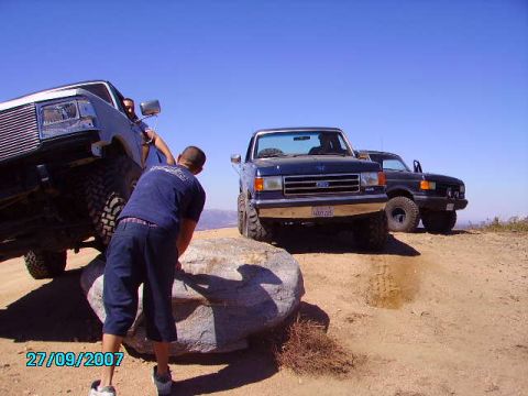 1990 Ford Bronco - Two Ford Broncos navigate a rocky off-road trail under a clear blue sky.