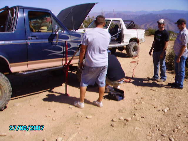 Two guys work on a blue Ford Bronco with flames, while another white pickup truck sits nearby on a dirt road with mountains in the background