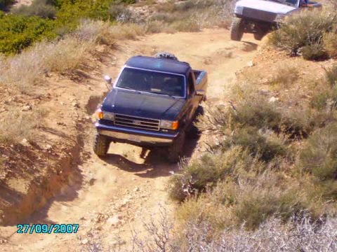 1990 Ford Bronco - A dark blue Ford F-Series truck navigates a dusty off-road trail with another vehicle visible in the background.