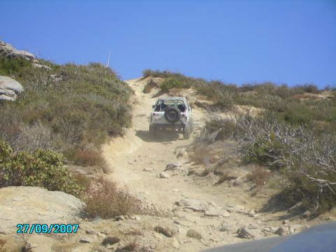 1990 Ford Bronco - A white SUV navigates a challenging off-road trail on a sunny day, kicking up dust as it ascends.