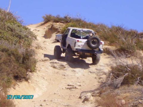 1990 Ford Bronco - Off-road SUV navigates a challenging dirt trail under a clear blue sky.