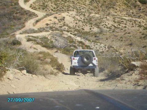 1990 Ford Bronco - White 1990 Ford Bronco navigates a winding dirt road in a dry, hilly terrain.
