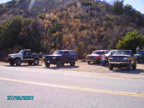 1990 Ford Bronco - A group of off-road vehicles are parked on the side of a road, with a dry, brush-covered hillside in the background.