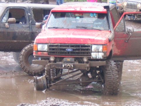 1989 Ford Bronco - A red off-road vehicle with its front passenger-side wheel detached is stuck in the mud.
