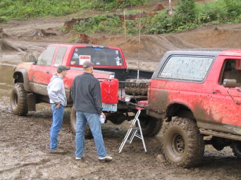 1989 Ford Bronco - Two red off-road vehicles, including a 1989 Ford Bronco, are stuck in the mud with two people observing.