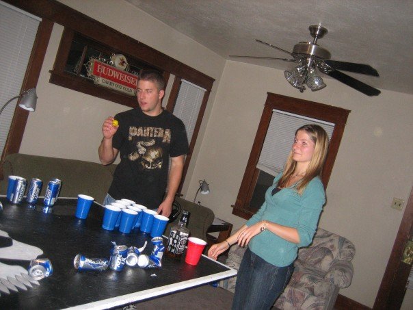 Man holding a ball, woman standing near a table with beer cups and cans set up for beer pong