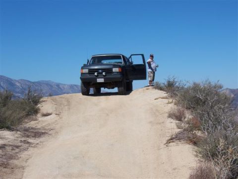 1995 Ford Bronco - A person stands by an off-road vehicle on a dusty trail with mountains in the background.