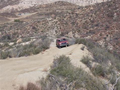 1995 Ford Bronco - A red 1995 Ford Bronco navigates a dusty, off-road trail in a dry, hilly terrain.