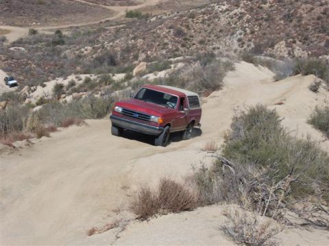 1995 Ford Bronco - Red Ford Bronco navigates a dusty, off-road trail in a dry, hilly landscape.