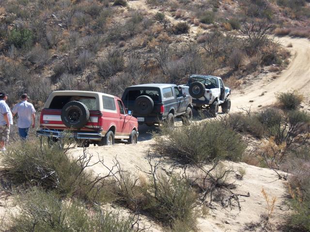 Three Ford Broncos driving on a dirt trail in a dry, hilly area. One red Bronco is in the foreground, followed by a dark Bronco, and then a white Bronco