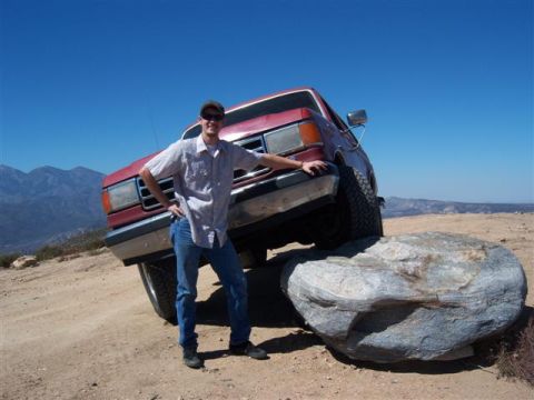 1995 Ford Bronco - A man poses with a red Ford Bronco that is tilted on a large rock on a dirt road with mountains in the background.