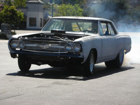 1998 Ford Crown Victoria - black-and-primer 1960s muscle car with a blower through the hood lays down a smoky burnout in a parking lot.